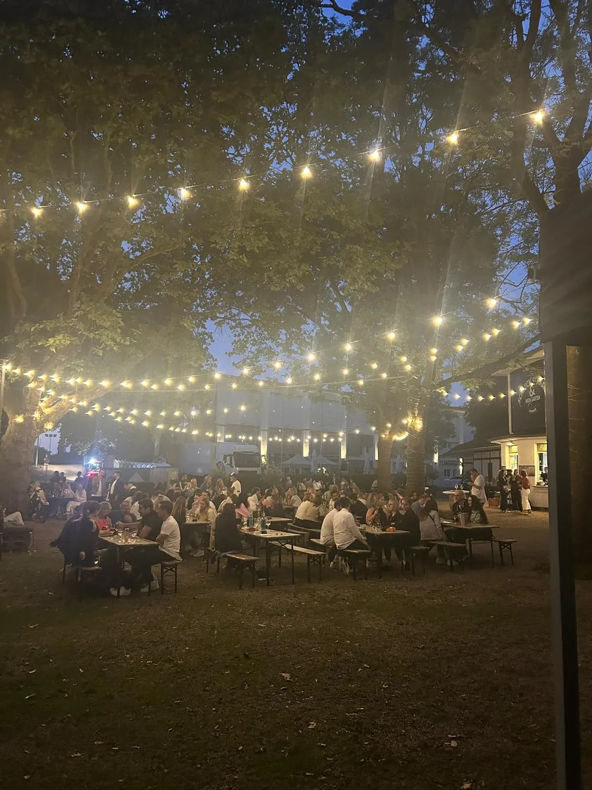 Der Garten von Schluck & Schnack an der Galopprennbahn am Abend, mit Lichterketten über langen Tischen